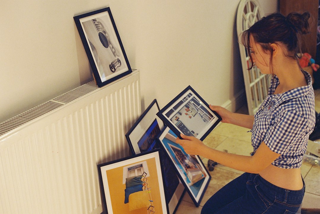A woman kneeling by a radiator and sorting through several framed pictures