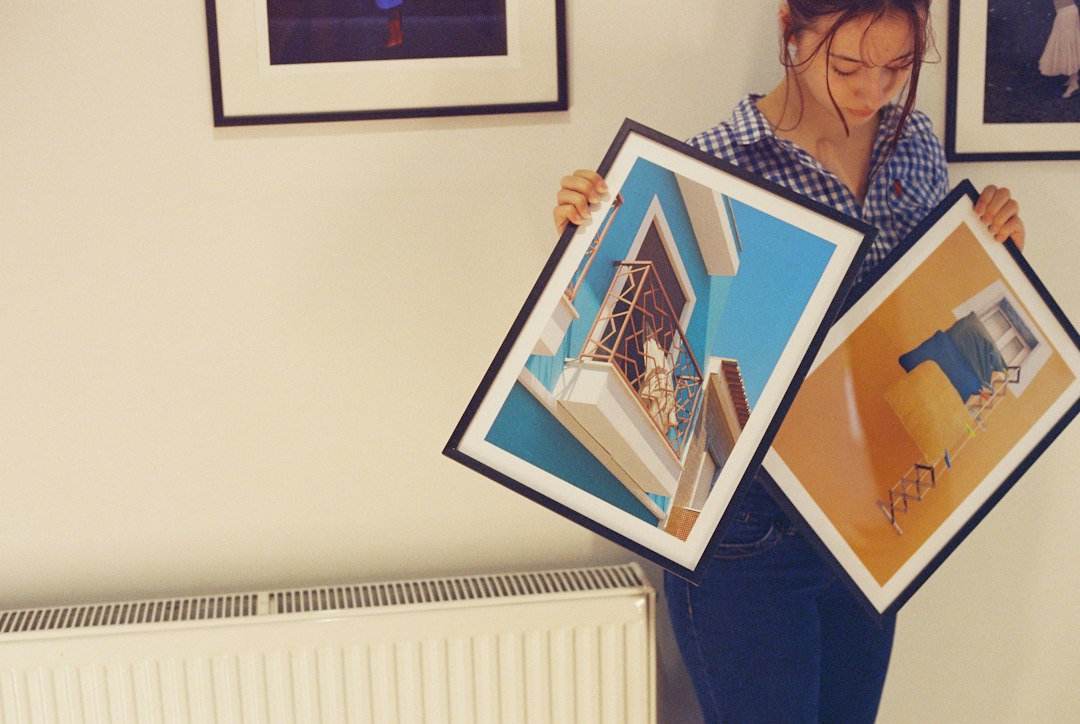 A woman holding two framed pictures against a wall with other framed artwork